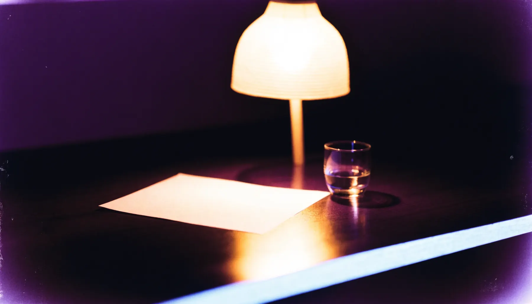 A single warm lamp lighting a blank sheet of paper on a dark desk